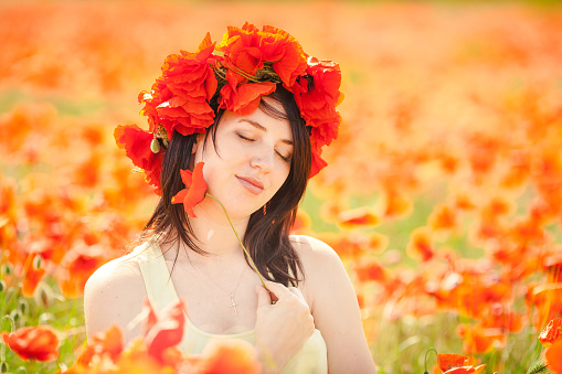 Portrait of young cheerful   woman with a poppy wreath on her head in a poppy field on sunny summer day. Portrait of a beautiful thoughtful girl walking in the fresh air, on a spring flower field. Walks in the countryside in a field of red poppies.