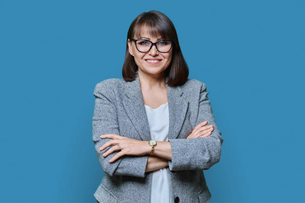 donna di mezza età positiva per gli affari con le braccia incrociate su priorità bassa blu - smiling women glasses assistance foto e immagini stock