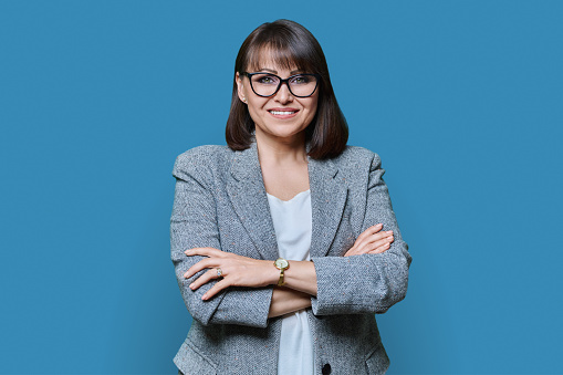 Business positive middle age woman with arms crossed on blue studio background. Confident woman with toothy smile in glasses jacket looking at camera. Business management leadership staff 40s people