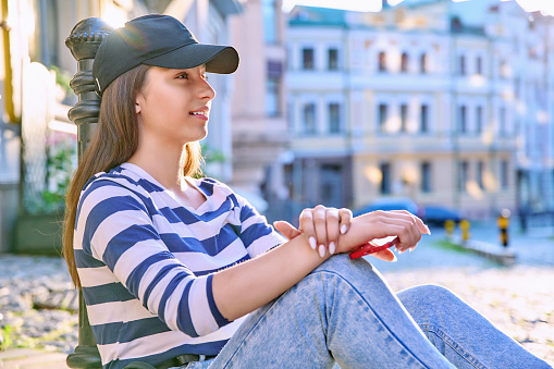 Beautiful teenage female sitting on the sidewalk. Fashionable female student in cap with backpack on city street. Adolescence, beauty, fashion, lifestyle, youth concept