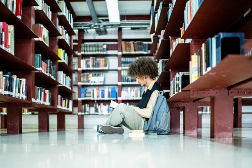 Young woman reading big old book and making a note while sitting in library in front of bookshelves. She wears green shirt and looks beautiful
