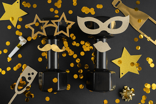 Two heavy dumbbells dressed up for New Year's Eve celebration, birthday party or carnival. Healthy fitness flat lay composition with golden decorations. Gym, workout Happy New Year resolution concept.