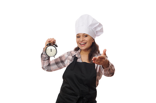 Charming female waitress in a chef's hat and black apron, holding an alarm clock in her hands and smiling, extends her hand to the camera, inviting to visit a new open catering establishment