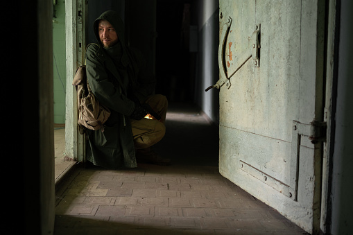 A dirty man in a hooded jacket is sitting near door in a large dark corridor, holding gun in his hands, looking into room. The concept of a survivor after the end of the world in search of food hiding