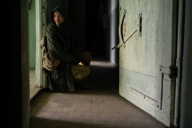dirty man in a hooded jacket is sitting near door in a large dark corridor, holding a gun in his hands, looking into room. concept of a survivor after the end of the world in search of food hiding. A dirty man in a hooded jacket is sitting near door in a large dark corridor, holding gun in his hands, looking into room. The concept of a survivor after the end of the world in search of food hiding hiding place stock pictures, royalty-free photos & images
