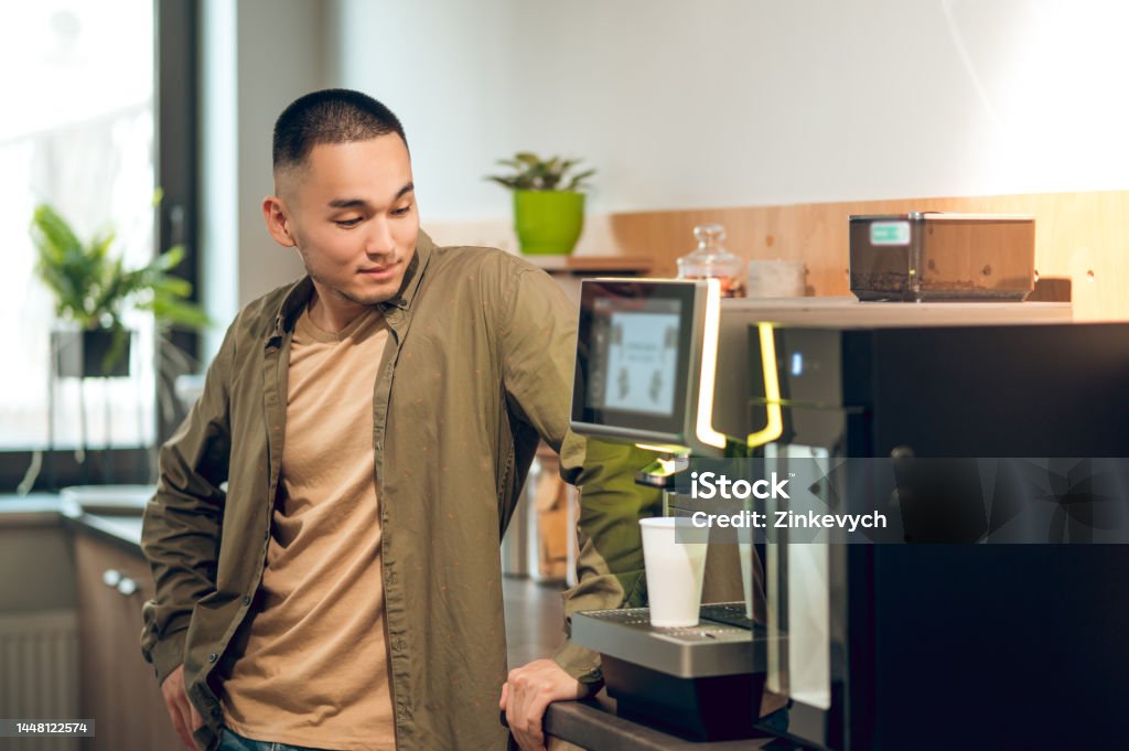 Company employee preparing a caffeinated beverage at work Young Asian corporate worker making himself cappuccino using the coffee machine in the office kitchen 25-29 Years Stock Photo