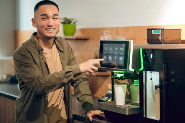 Waist-up portrait of a smiling cheerful young man tapping on the coffee machine touch screen and choosing the drink type