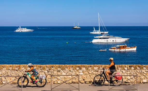 Tourists cycling on promenade in front of harbor and yachts with French riviera seascape in Antibes resort town in France stock photo
