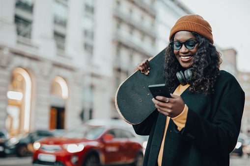 A young woman, a young black woman with a skateboard in her hands walks down the street of Barcelona, dressed casually, with headphones around her neck and a knitted winter hat, sightseeing in Barcelona and using a mobile phone
