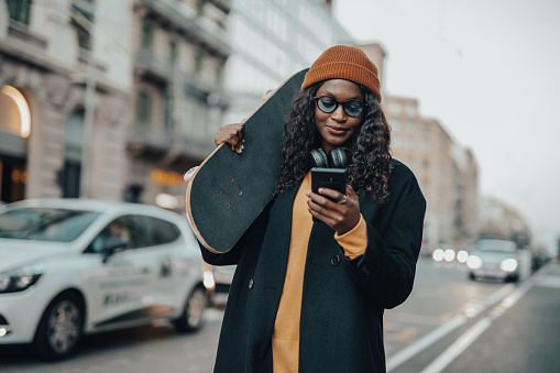A young woman, a young black woman with a skateboard in her hands walks down the street of Barcelona, dressed casually, with headphones around her neck and a knitted winter hat, sightseeing in Barcelona and using a mobile phone