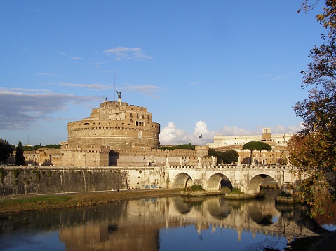 A castle, buildings and bridge across a river in Rome. The buildings and blue sky reflect on the water's surface.