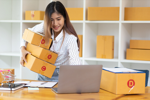 Startup SME small business entrepreneur of freelance Asian woman using laptop and box to receive and review orders online to prepare to pack sell to customers, online sme business ideas.