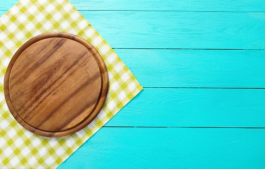 Frame of round cutting board for pizza  food and green plaid tablecloth. Blue wooden background. Top view mockup. Copy space