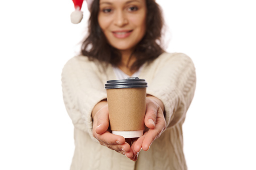 Selective focus on a takeaway hot drink in disposable cardboard cup in the hands of a blurred woman in Santa hat, smiling on camera, isolated over white background. Beverage to go. Mockup. Copy space.