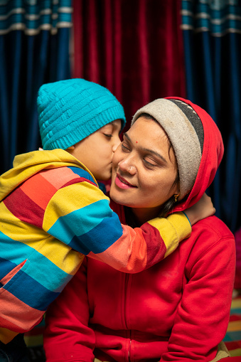 Winter portrait of a happy, affectionate son embracing and kissing his mother on the cheeks. They are sitting together at home and wearing warm clothes in winter.