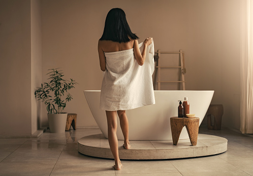Back view of slim woman in towel going to take bath in white bathtub in luxury bathroom