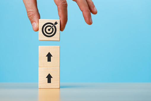 Hand putting stack of wooden blocks with arrow and target icons on a blue background copy space. Business development strategy, advancement and goal concept.