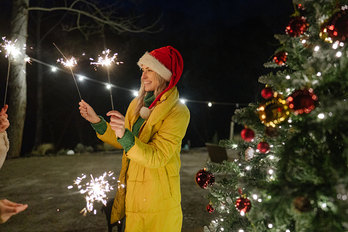 Photo of a young woman by the Christmas tree, holding sparklers and having a great time.