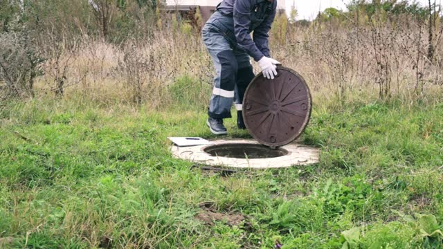 a male plumber in overalls opens the manhole of a water well with a crowbar. Inspection of water meter readings