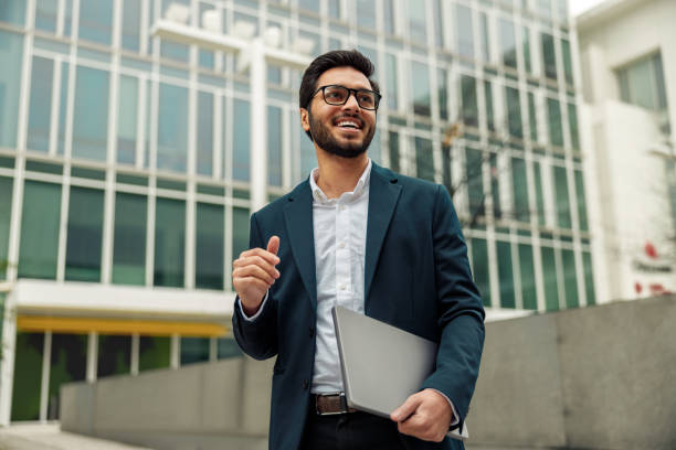 hombre de negocios indio sonriente con traje y gafas con computadora portátil cerca del edificio de oficinas - people clothing elegance built structure fotografías e imágenes de stock