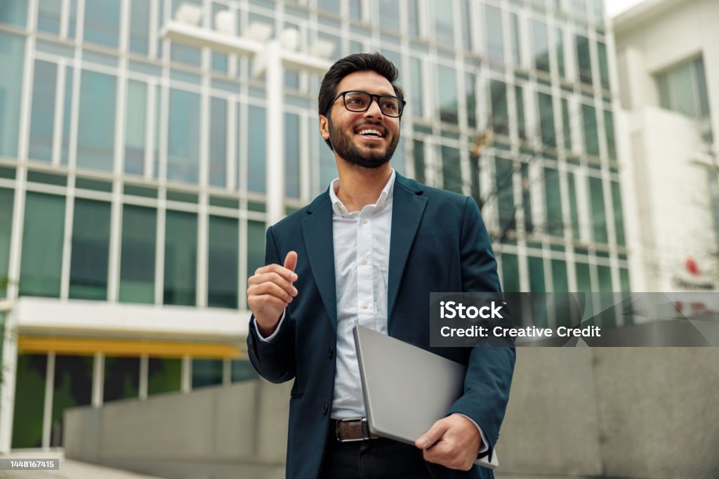 Smiling indian businessman in suit and glasses with laptop near office building Smiling businessman in suit with laptop near modern office building is looking at side Businessman Stock Photo