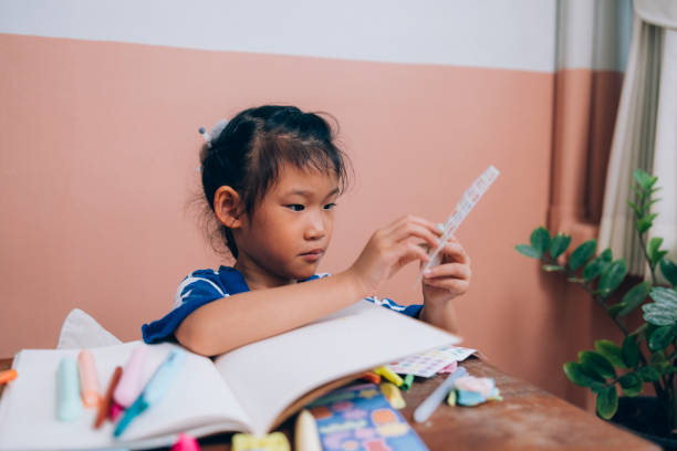 Asian Girl Focused On Her Stickers Little Asian girl sitting at a small desk at home, enjoying her hobby of drawing and colouring and using stickers. kid stickers stock pictures, royalty-free photos & images