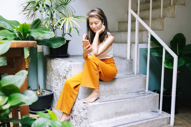 A Happy Beautiful Woman In White T-Shirt And Orange Trousers, Barefoot, Watching Something Online On Her Mobile Phone While Sitting In The Corridor Of Her Building stock photo