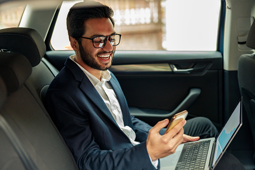 Smiling businessman in suit working laptop while riding in car to office. High quality photo