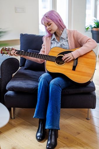 A young woman is sitting on a couch in a living room in a modern house and playing an acoustic guitar.