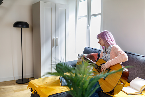A young woman is sitting on a couch in a living room in a modern house and playing an acoustic guitar with online help using a laptop.