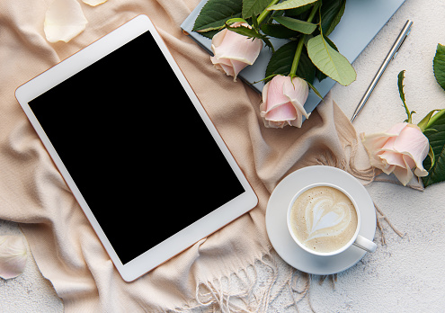 Minimalist workspace with digital tablet empty screen mockup, cozy scarf, a cup of coffee and roses flowers on concrete background. Top view, flat lay