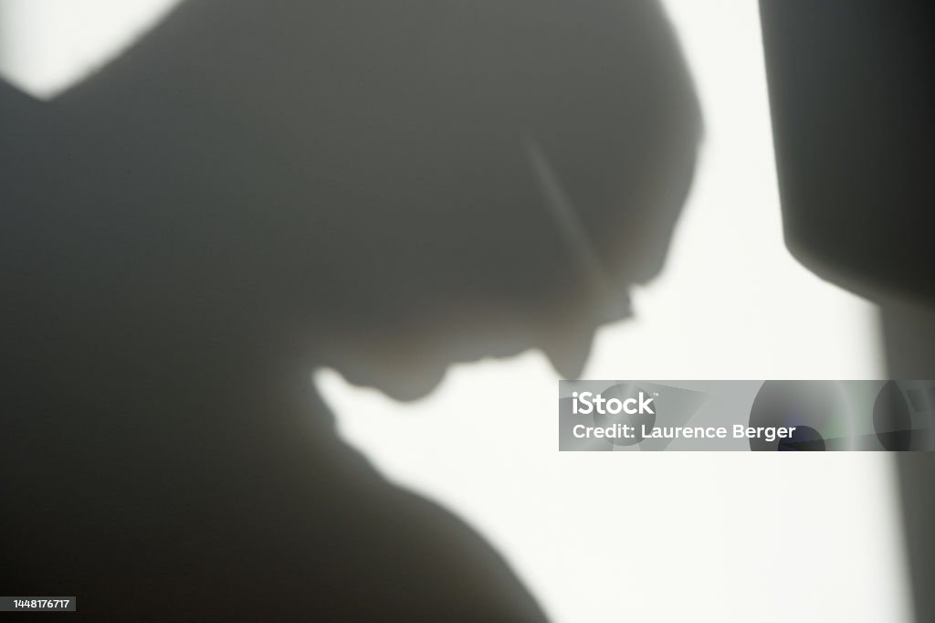 Sunlit shadow of a mature adult male head bowed Dramatic sunlit shadow of a mature  adult male head bowed in silhouette. Plain background.  Illustrates sadness, depression despair or defeat Adult Stock Photo