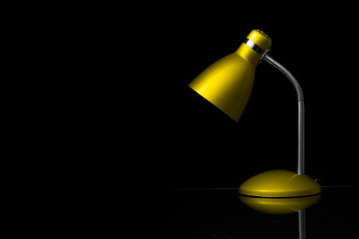 Yellow desk lamp on black background, with reflection on the table. Copy space. Horizontal orientation. No people.