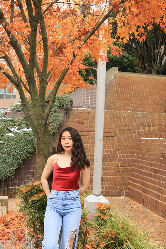 A Chinese model in an Urban garden with trees if fall foliage, and a bit of snow on the ground. The model is wearing a red sleeveless top, torn, blue jeans and makeup.