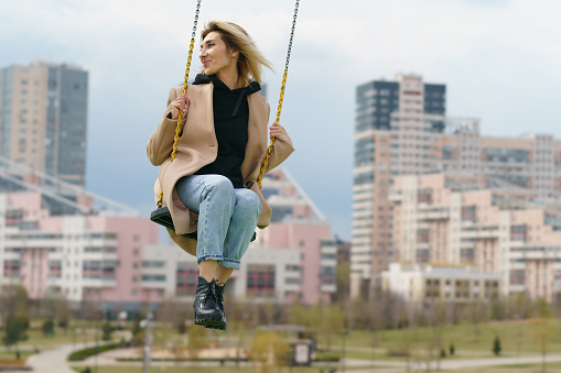 Funny young female model sits on the adults' big swing on the Moscow city street. Spring day. Leisure time for relaxation. Moscow cityscape as background.