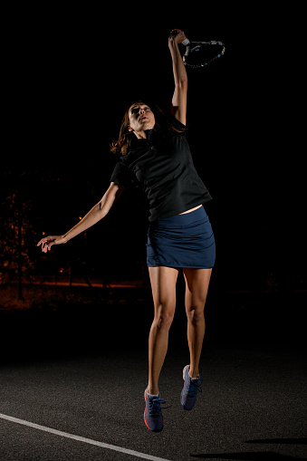 female tennis player with tennis racket in her hand bouncing to hit the tennis ball. Dark background. Movement, sport, healthy lifestyle concept.
