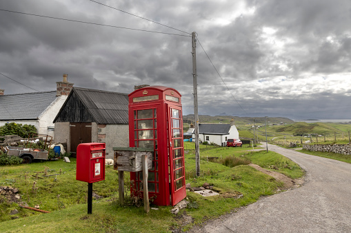 British vintage red phone booth in the middle of nowhere, along a country road in Scotland. overcast cloudy day, rain drizzle. Contrast between technology and nature, red and green.