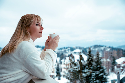 Blonde Woman in bathrobe with morning coffee on balcony with winter landscape view in snowy mountains. Pretty female on winter ski vacation. Copy space
