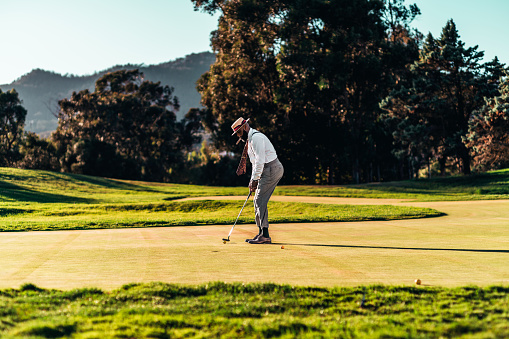 A fashionable handsome bearded black senior with a cigar, in straw hat and a tailored outfit standing on lush green golf course with a putting club in his hand aiming the yellow ball to the hole