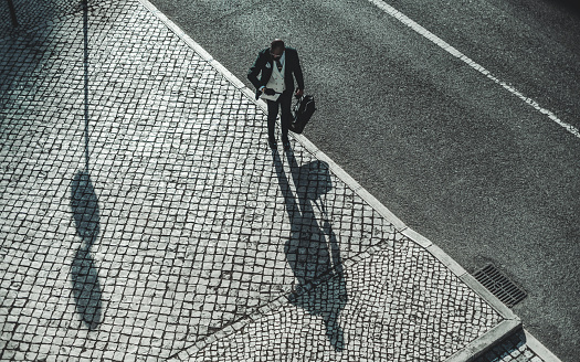 Drone view of a bald black man, dressed in a tie vest, on the edge of the sidewalk where you see a shadow of him and a traffic sign, his hands loaded with a suitcase and a newspaper