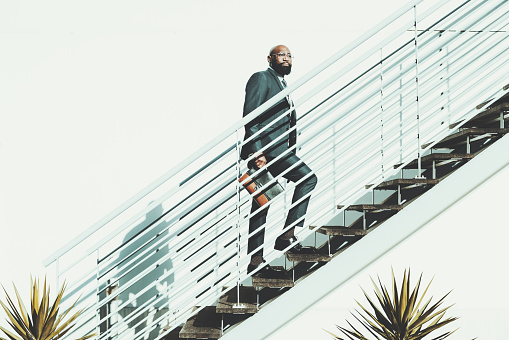 A bald black bearded man walks up the stairs. The African male is ascending a white metallic staircase on a shiny day, wears a dark suit and holds a leather purse in a very clean and cactus aesthetic