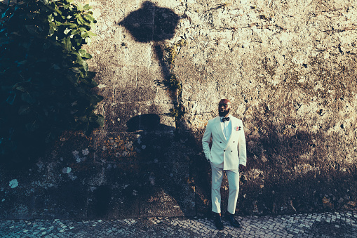 Tall African bald and bearded man dressed in a white suit with a polka-dotted bow, glasses and matching shoes. Behind him a brown stoned wall with a shadow of street lamp and a climbing vine