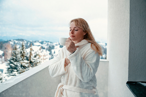 Blonde Woman in bathrobe with coffee on hotel balcony with winter landscape view in snowy mountains. Copy space