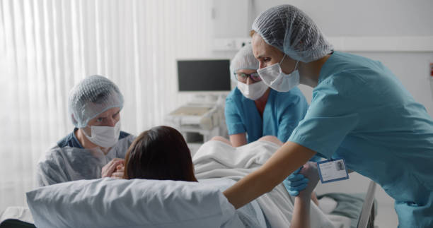 Woman giving birth with husband holds her hand in support and obstetricians assisting. Woman giving birth with husband holds her hand in support and obstetricians assisting. Back view of medical staff in protective uniform helping pregnant woman in labor childbirth stock pictures, royalty-free photos & images