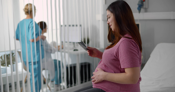 Young pregnant woman looking at ultrasound image sitting at bed in hospital. Smiling expectant female holding sonogram picture after examination in clinic