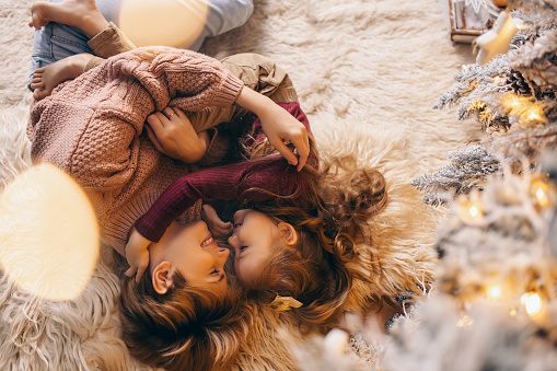 A young mother and daughter 5-6 years old in casual clothes lie on a soft carpet in a cozy, and decorated Christmas living room at home against the backdrop of a Christmas tree.