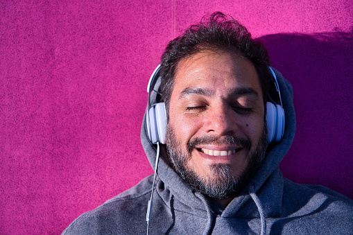 Portrait of a smiling latin man listening to relaxed music with his eyes closed, wearing white headphones on a vivid magenta background