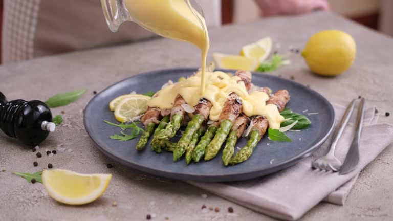 pouring hollandaise sauce onto freshly cooked asparagus wrapped with bacon on ceramic plate