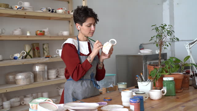 Young creative talented French female ceramist in apron painting mug, standing behind table in studio