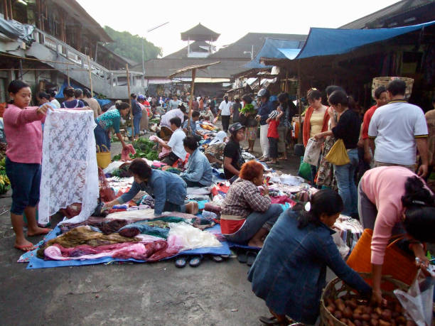Horizontal closeup photo of a busy morning market scene with a crowd of people looking at traditional Balinese fabrics, secondhand clothing and a variety of household items on tarpaulins on the ground or displayed in covered outdoor market stalls. Ubud, Bali. 1st March, 2008.